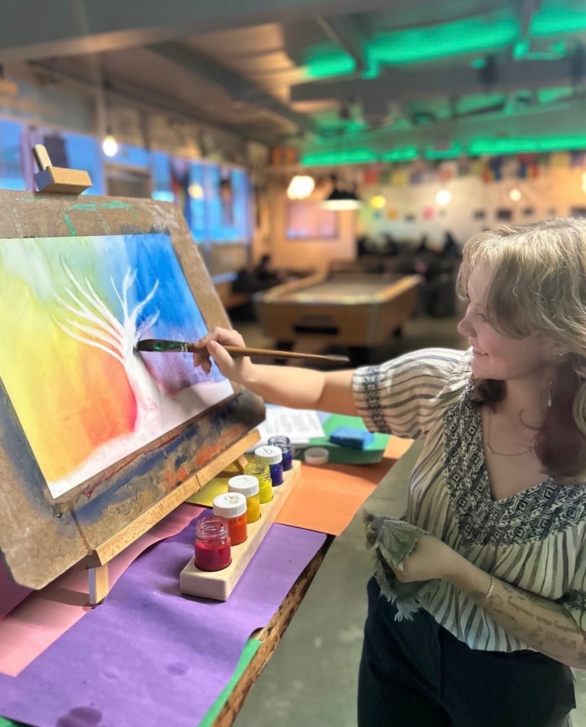 An artist painting a white tree over a rainbow-colored background at an easel