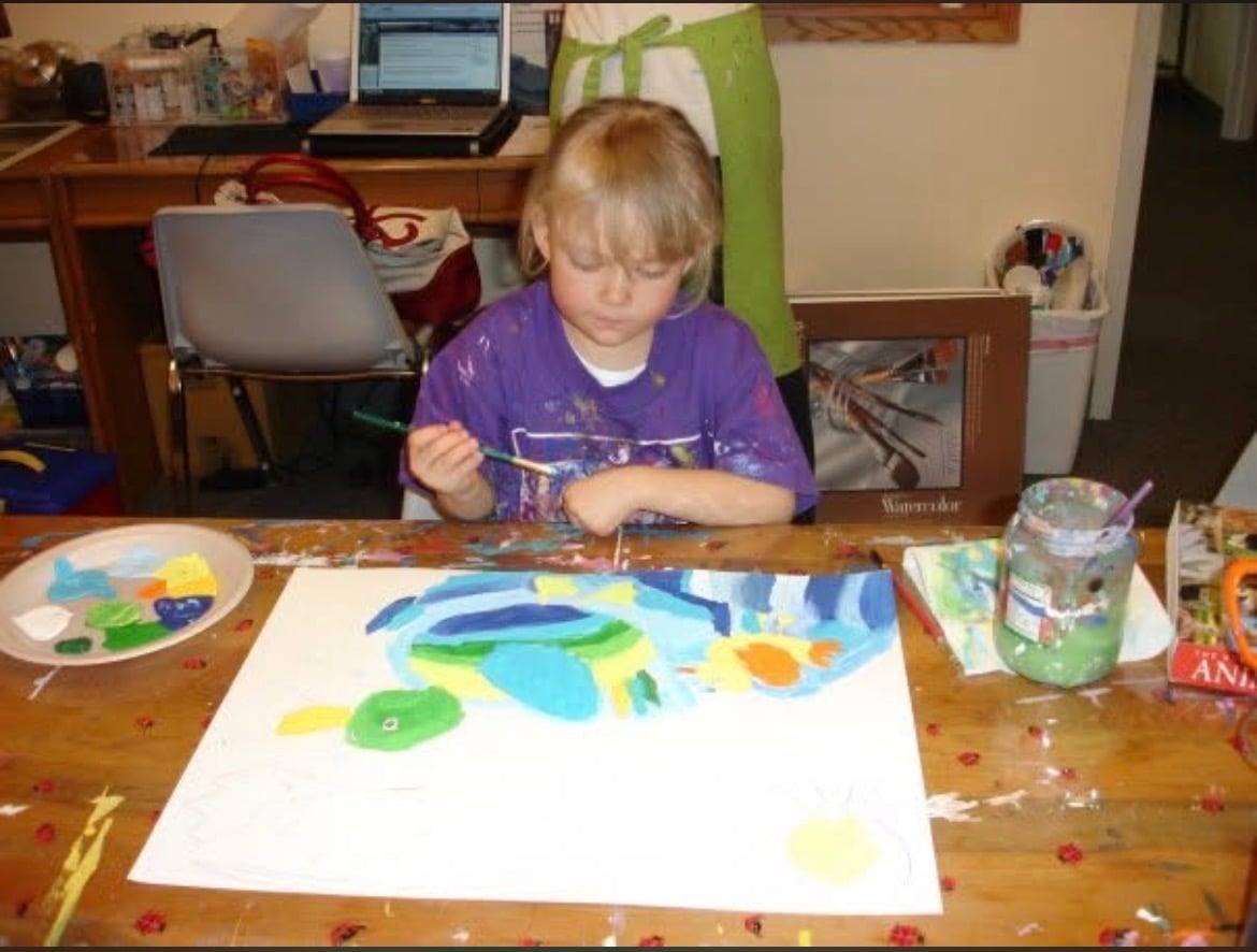 A child in a purple shirt painting a colorful bird at a table